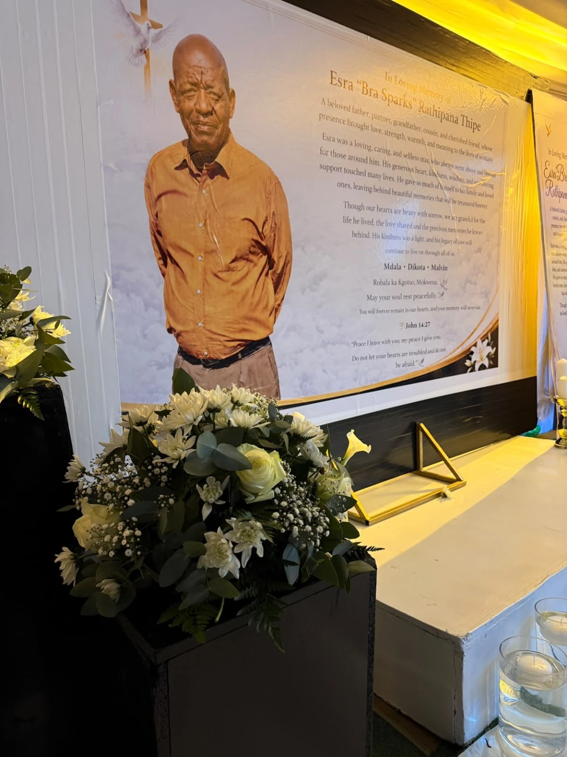 A close-up of a memorial backdrop, framed with cream flowers, greenery and warm gold ambient light.